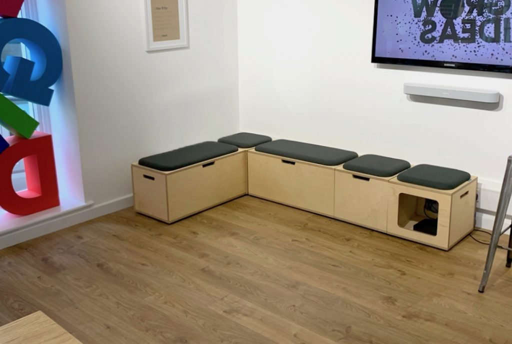 Office reception breakout space, with wooden plywood storage benches, with upholstered seat pads in grey fabric